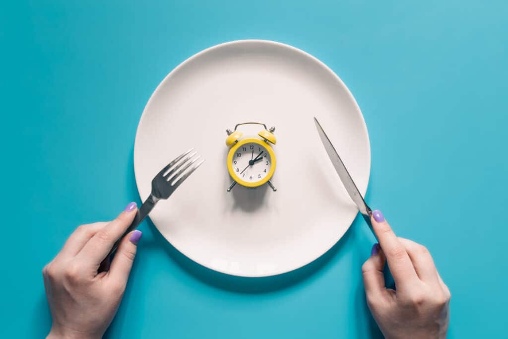 Hands holding knife and fork above alarm clock on a plate on blue background.