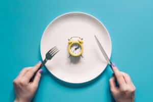 Hands holding knife and fork above alarm clock on a plate on blue background.